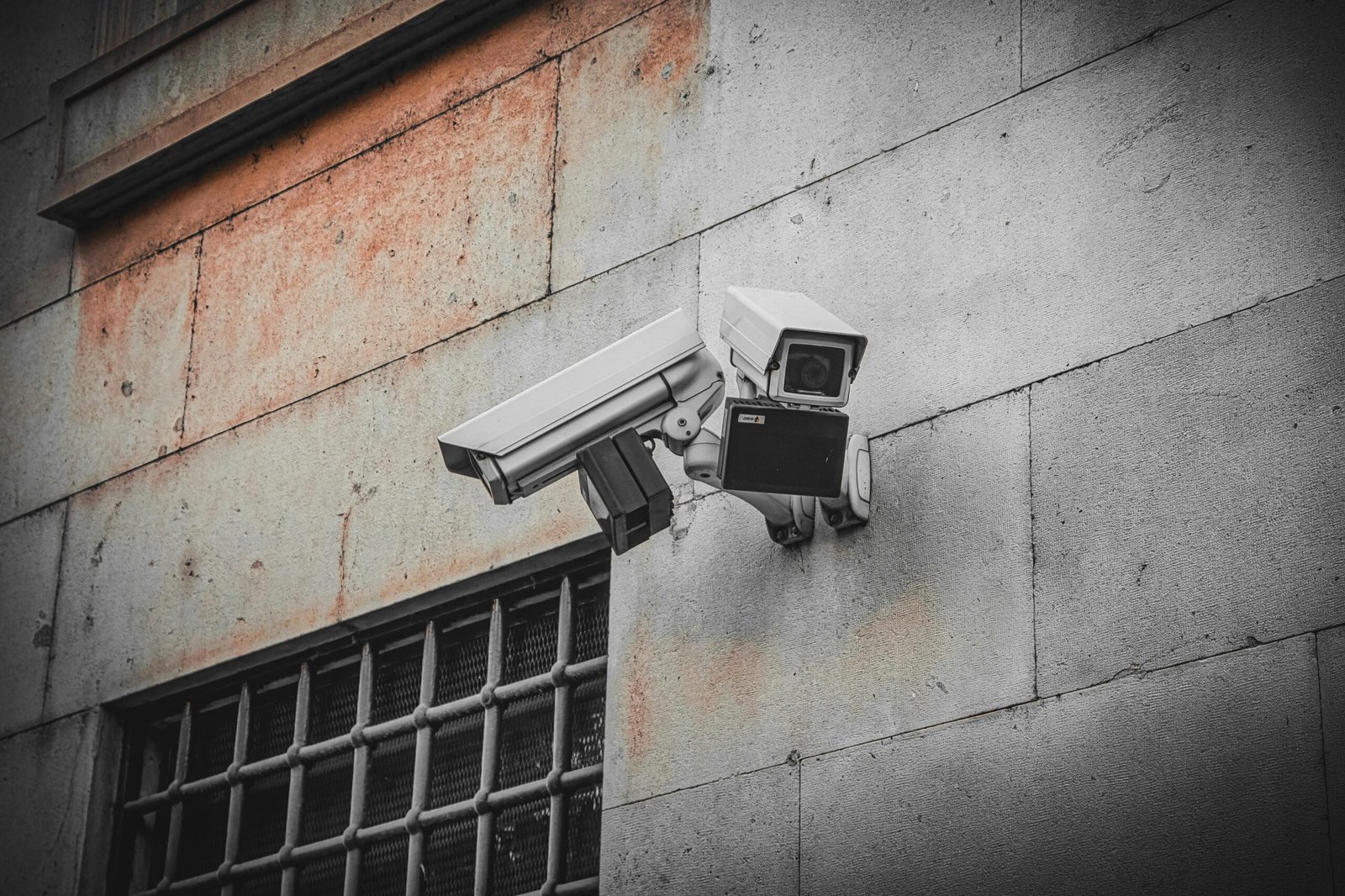Two surveillance cameras mounted on an aged concrete wall beside a barred window, capturing urban security.